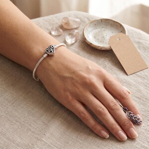 May include: A silver snake chain bracelet featuring a heart-shaped charm with an intricate tree of life cutout design. The charm is centered on the flexible metal band, which has a polished finish and a secure clasp mechanism for closure.