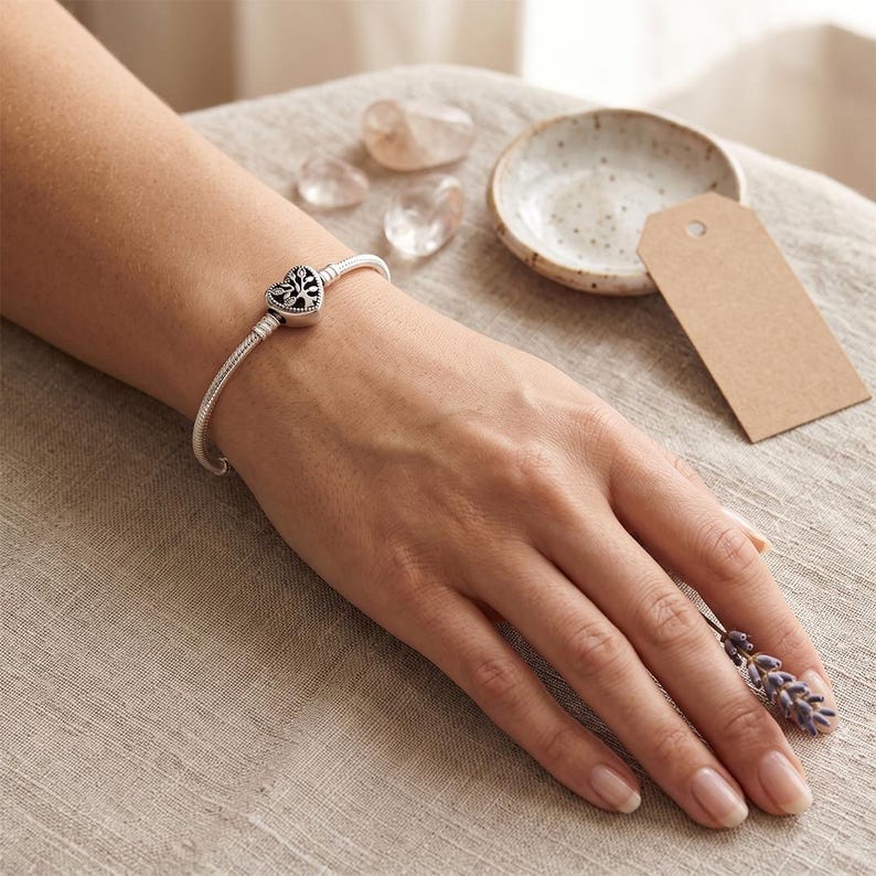 May include: A silver snake chain bracelet featuring a heart-shaped charm with an intricate tree of life cutout design. The charm is centered on the flexible metal band, which has a polished finish and a secure clasp mechanism for closure.
