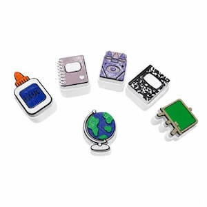 May include: A set of six school-themed charms including a blue glue bottle with the word GLUE, a purple spiral notebook, a purple book with an eye design, a black and white composition notebook, a green chalkboard, and a blue and green globe.