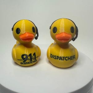 May include: Two yellow duck figurines with orange beaks and black headsets. One duck has "911 DISPATCHER" printed on its chest, the other has "DISPATCHER" printed on its chest. The ducks are on a white surface.
