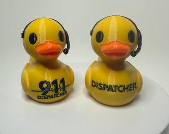 Könnte beinhalten: Zwei gelbe Entenfiguren mit orangefarbenen Schnäbeln und schwarzen Headsets. Eine Ente hat "911 DISPATCHER" auf der Brust stehen, die andere "DISPATCHER". Die Enten stehen auf einer weißen Oberfläche.