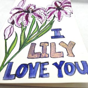 May include: A handmade greeting card featuring watercolor illustrations of purple and white speckled lilies with green leaves. The card reads "I Lily Love You" in blue and gold lettering, perfect for a floral gift.