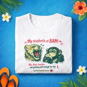 May include: White t-shirt with a humorous graphic. The design features a roaring lion and a contemplative monkey, with text that reads "My students at 8AM" and "Me, their teacher, not getting paid enough for this." The shirt also says "Certified Rage Sait Survivor."