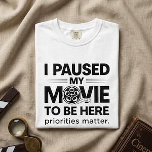 Op de afbeelding: Wit T-shirt met de zwarte tekst "I PAUSED MY MOVIE TO BE HERE priorities matter." en een filmrol-afbeelding. Het shirt is opgevouwen op een beige stoffen ondergrond, met een klapbord en een vergrootglas.