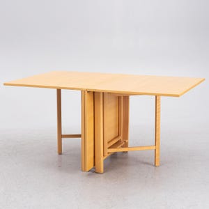 May include: A light-coloured wooden drop-leaf table with a rectangular top and four legs. The table is partially folded, showing the folding mechanism. The wood has a natural finish.