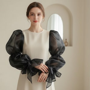 May include: A cream-colored dress with black, sheer, puffy sleeves. The sleeves are gathered at the wrist and adorned with large bows. The dress has a simple, round neckline and a fitted bodice.