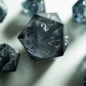 May include: Close-up of several polyhedral dice with a dark, translucent appearance. The dice have silver numbering and a speckled, glittery interior. The dice are arranged on a white surface, with shadows adding depth.