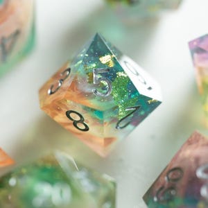 May include: Close-up of several polyhedral dice with a colorful, layered design. The dice are transparent with embedded glitter and a mix of orange, green, and blue hues. Each die has silver numbering.