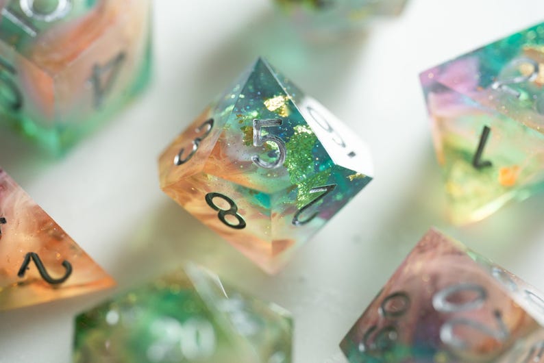 May include: Close-up of several polyhedral dice with a colorful, layered design. The dice are transparent with embedded glitter and a mix of orange, green, and blue hues. Each die has silver numbering.