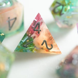 May include: Close-up of colorful resin dice with embedded floral elements. The dice are in various shapes, including a triangular D4 die with the number 4 and a D6 die with the number 5. The dice have a translucent appearance with a mix of pink, orange, green, and gold.