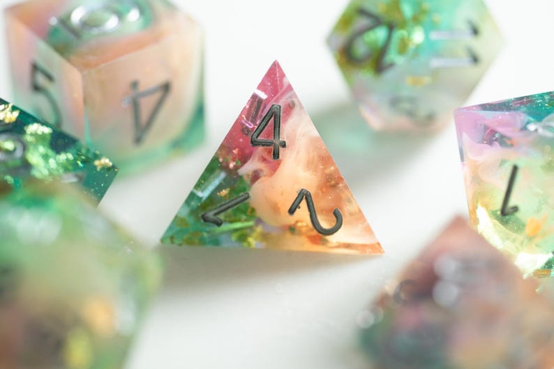 May include: Close-up of colorful resin dice with embedded floral elements. The dice are in various shapes, including a triangular D4 die with the number 4 and a D6 die with the number 5. The dice have a translucent appearance with a mix of pink, orange, green, and gold.