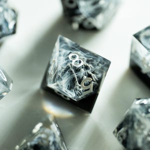 Könnte beinhalten: Eine Sammlung von polyedrischen Würfeln mit einem marmorierten Schwarz-Weiß-Design. Die Würfel sind transparent mit weißen Zahlen und einem Wirbelmuster. Die Würfel sind auf einer weißen Oberfläche angeordnet, wobei einige fokussiert und andere unscharf sind.