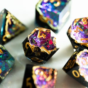 May include: A set of seven polyhedral dice for tabletop games. The dice are black with gold numbering and filled with purple, blue, and pink glitter. These resin dice are designed for role-playing games.