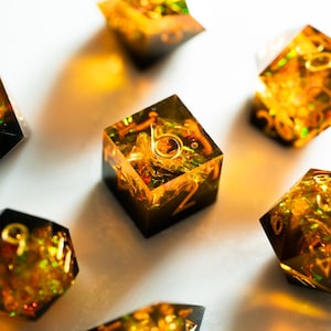 May include: A collection of polyhedral dice with a unique design. The dice are a mix of black and gold, with gold numbering. The interior of the dice has a golden, glittery appearance. The dice are scattered on a white surface.