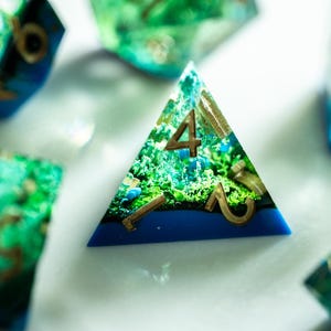 Peut inclure: Gros plan sur un ensemble de dés polyédriques. Le dé D4 est bleu et vert avec des chiffres dorés. Les dés ont un motif de paysage à l'intérieur. D'autres dés sont visibles en arrière-plan.