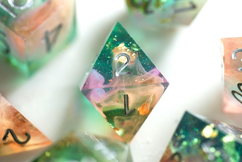 May include: Close-up of a set of handmade resin dice. The dice are clear with colorful swirls of pink, orange, and green. The number 2 is visible on the face of one die, with a silver metallic finish. Gold flakes are suspended within the resin.