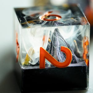 May include: A close-up of a unique, handmade resin die. The die features an orange number 2, set against a backdrop of a mountain range and wispy white and gray smoke-like patterns. The die is encased in a clear cube.