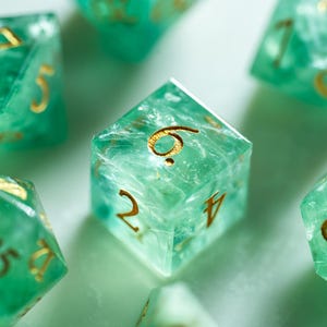 May include: Close-up of several translucent green dice with gold-colored numbers. The dice are scattered across a light surface. The dice are a light green color with gold numbers, perfect for tabletop games.