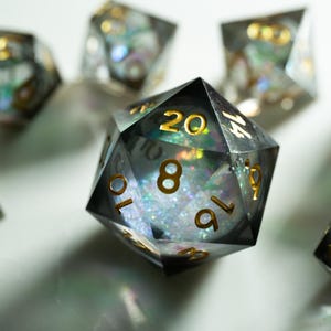 May include: A collection of polyhedral dice with a clear, iridescent interior and gold-colored numbering. The dice are designed for tabletop gaming, with a focus on the twenty-sided die in the foreground. The dice are set against a white background.