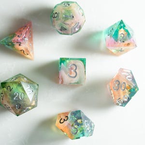 May include: A collection of colorful, handmade resin dice in various geometric shapes. The dice feature a mix of pastel colors, including pink, green, and orange, with silver numbering. Ideal for tabletop gaming.