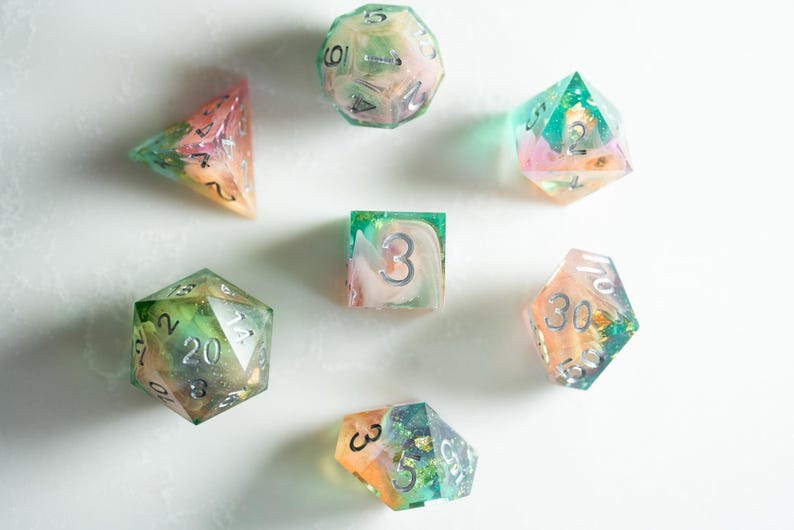 May include: A collection of colorful, handmade resin dice in various geometric shapes. The dice feature a mix of pastel colors, including pink, green, and orange, with silver numbering. Ideal for tabletop gaming.