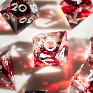 May include: Close-up of a set of polyhedral dice. The dice are clear with red and gold inclusions, and white numbering. The dice are in various shapes, including a d20, d6, and d4, and are scattered on a white surface.