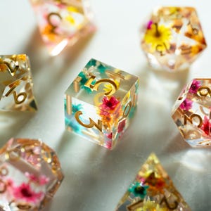 May include: Seven clear resin dice with embedded dried flowers in various shapes, including a cube and a pyramid. The dice feature gold numbering and are filled with colourful flowers, such as yellow, pink, and green. The dice are for tabletop games.
