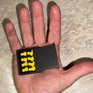 Puede incluir: Un estuche rectangular negro con tapa texturizada, abierto para revelar tapones para los oídos amarillos. El estuche se sostiene en la palma de la mano abierta. Los tapones para los oídos están en un patrón de cuadrícula.