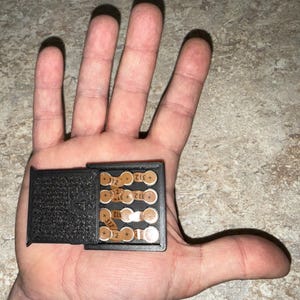 Puede incluir: Una caja negra rectangular con bisagras, probablemente impresa en 3D, que contiene varias pilas pequeñas, redondas y de color cobre. Cada pila tiene el número "+312+" impreso. La caja está abierta, revelando las pilas en su interior.