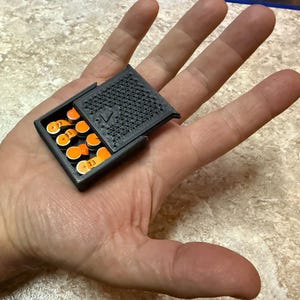 以下が含まれることがあります： テクスチャード加工された蓋が付いた、小さな黒いヒンジ付き容器。オレンジと白のゲームピースがいくつか入っています。ピースには「+13」の数字が印刷されています。容器は開いており、中身が見えています。