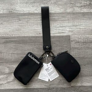 May include: Black dual pouch wristlet with a black strap and silver-tone hardware. One pouch has the word "lululemon" printed in white. The wristlet includes a tag with product information.