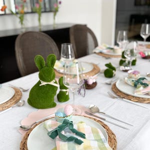 May include: Easter table setting with green bunny figurines, pastel-colored napkins, and decorative eggs. The table is set with white plates, woven placemats, and silverware. Wine glasses and floral arrangements complete the festive look.