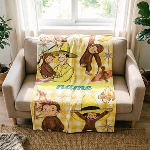 Pode incluir: Uma manta de lã xadrez amarela e branca com Curious George e o homem do chapéu amarelo. O design inclui ilustrações das personagens, uma fogueira e uma cadeira de nadador-salvador, com a palavra name impressa em azul.