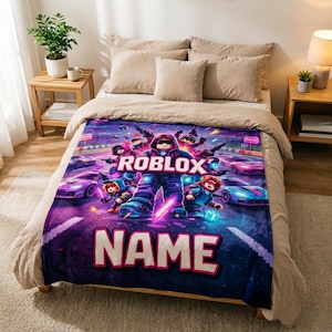 Puede incluir: Una manta de felpa con un gr&aacute;fico vibrante de personajes de Roblox y coches de carreras en tonos morados, azules y rosas. La palabra "ROBLOX" se muestra de forma destacada, con la palabra "NAME" debajo. La manta est&aacute; sobre una cama.