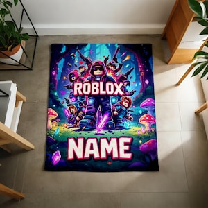 Puede incluir: Una manta rectangular con un dise&ntilde;o vibrante con tem&aacute;tica de Roblox. La manta muestra un grupo de personajes de Roblox con la palabra "ROBLOX" en un lugar destacado. Debajo, la palabra "NAME" est&aacute; escrita en letras grandes, lo que sugiere personalizaci&oacute;n.
