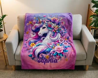 Puede incluir: Una manta de felpa morada con un diseño de unicornio colorido. El unicornio tiene un cuerpo blanco, un cuerno dorado y una melena y cola fluidas en tonos de rosa, azul y morado. El nombre "Sophia" está impreso en la manta.