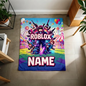 Puede incluir: Una manta rectangular azul con un dise&ntilde;o colorido de Roblox. La manta muestra el logotipo de Roblox y la palabra "NAME" en letras grandes. El dise&ntilde;o incluye personajes de dibujos animados, una noria y globos.