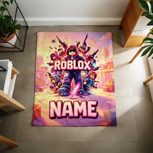 Puede incluir: Una manta rectangular con una vibrante ilustraci&oacute;n de personajes de Roblox en un entorno urbano. La palabra "ROBLOX" se muestra de forma destacada, con el nombre "NAME" debajo. La manta tiene un dise&ntilde;o colorido con tonos de rosa, morado y azul.