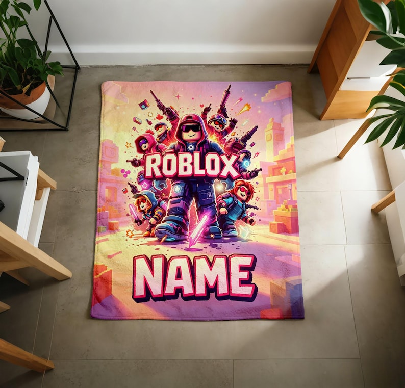 Puede incluir: Una manta rectangular con una vibrante ilustraci&oacute;n de personajes de Roblox en un entorno urbano. La palabra "ROBLOX" se muestra de forma destacada, con el nombre "NAME" debajo. La manta tiene un dise&ntilde;o colorido con tonos de rosa, morado y azul.