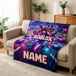 Puede incluir: Una manta de felpa vibrante con un dise&ntilde;o tem&aacute;tico de Roblox. La manta muestra un grupo de personajes de Roblox en una escena de carreras, con la palabra "ROBLOX" en un lugar destacado. La parte inferior de la manta tiene espacio para un nombre.