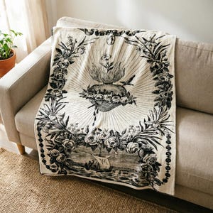 May include: Cream-colored throw blanket with a black illustration of a heart, flowers, and a ship. The blanket is draped over a beige sofa. The design is framed by a floral border.