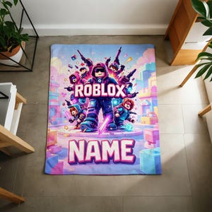Puede incluir: Una colorida manta con un dise&ntilde;o tem&aacute;tico de Roblox. La manta muestra la palabra "ROBLOX" en letras grandes y audaces, junto con varios personajes de dibujos animados. La palabra "NAME" tambi&eacute;n se muestra de forma destacada. La manta tiene una combinaci&oacute;n de colores azul, rosa y morado.