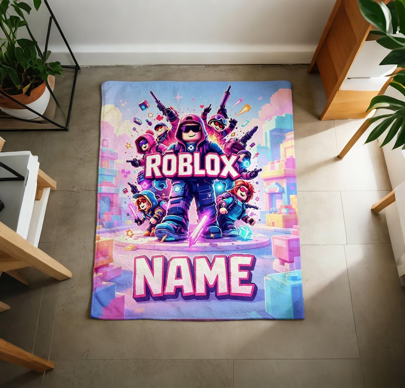 Puede incluir: Una colorida manta con un dise&ntilde;o tem&aacute;tico de Roblox. La manta muestra la palabra "ROBLOX" en letras grandes y audaces, junto con varios personajes de dibujos animados. La palabra "NAME" tambi&eacute;n se muestra de forma destacada. La manta tiene una combinaci&oacute;n de colores azul, rosa y morado.