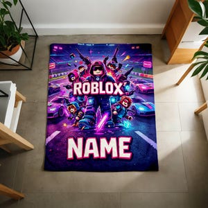 Puede incluir: Una manta rectangular con un dise&ntilde;o vibrante con tem&aacute;tica de Roblox. La manta muestra un personaje central de Roblox rodeado de otros personajes y coches de carreras. Las palabras "ROBLOX" y la palabra personalizable "NAME" se muestran de forma destacada.
