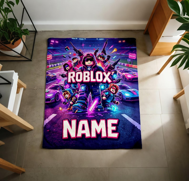 Puede incluir: Una manta rectangular con un dise&ntilde;o vibrante con tem&aacute;tica de Roblox. La manta muestra un personaje central de Roblox rodeado de otros personajes y coches de carreras. Las palabras "ROBLOX" y la palabra personalizable "NAME" se muestran de forma destacada.