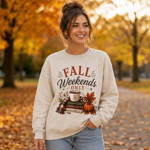 Puede incluir: Una sudadera de cuello redondo color crema con un estampado gráfico con el texto "FALL WEEKENDS ONLY" sobre una ilustración de una taza de café, una pila de libros etiquetados "PUMPKIN SPICE" y "CRUNCHY LEAVES", una bufanda de cuadros y una calabaza.