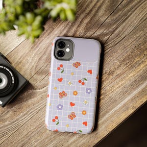 May include: A lavender phone case with a grid pattern, adorned with red hearts, cherries, butterflies, daisies, and stars. The case has a camera cutout and a solid lavender top section. The case is on a wooden surface.