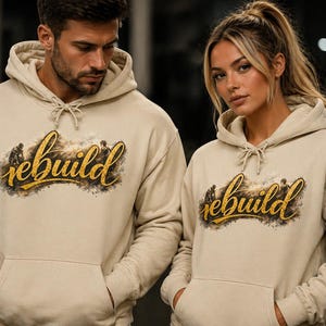 Puede incluir: Dos personas con sudaderas con capucha beige a juego. Las sudaderas tienen la palabra "rebuild" en letras doradas y un diseño de silueta. Tienen cordones y un bolsillo delantero, para deporte o uso diario.