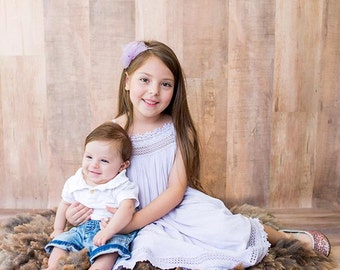 May include: Two young girls, one wearing a white shirt and blue denim shorts, and the other wearing a lavender dress, are sitting on a brown fluffy rug. The older girl is holding the younger girl. The background is a wood-paneled wall. The text "Photography by Anna Frances" is written in white at the bottom of the image.