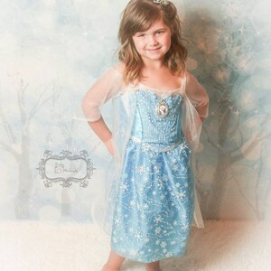 May include: A young girl wearing a blue and white snowflake patterned dress with a sheer overlay and a silver tiara. She is standing on a white fluffy surface.